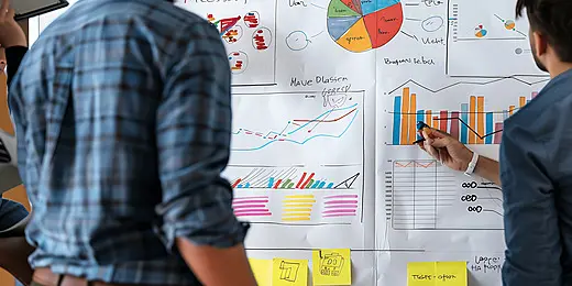 Two people discuss SEO strategies and website plans at a whiteboard filled with graphs, charts, and colorful sticky notes.