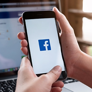Person holding a smartphone with Facebook open, blurred laptop behind, illustrating digital marketing and web design.