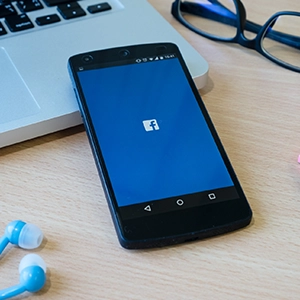Smartphone with Facebook login on a desk alongside laptop, glasses, and earphones—tools for web design and SEO work.