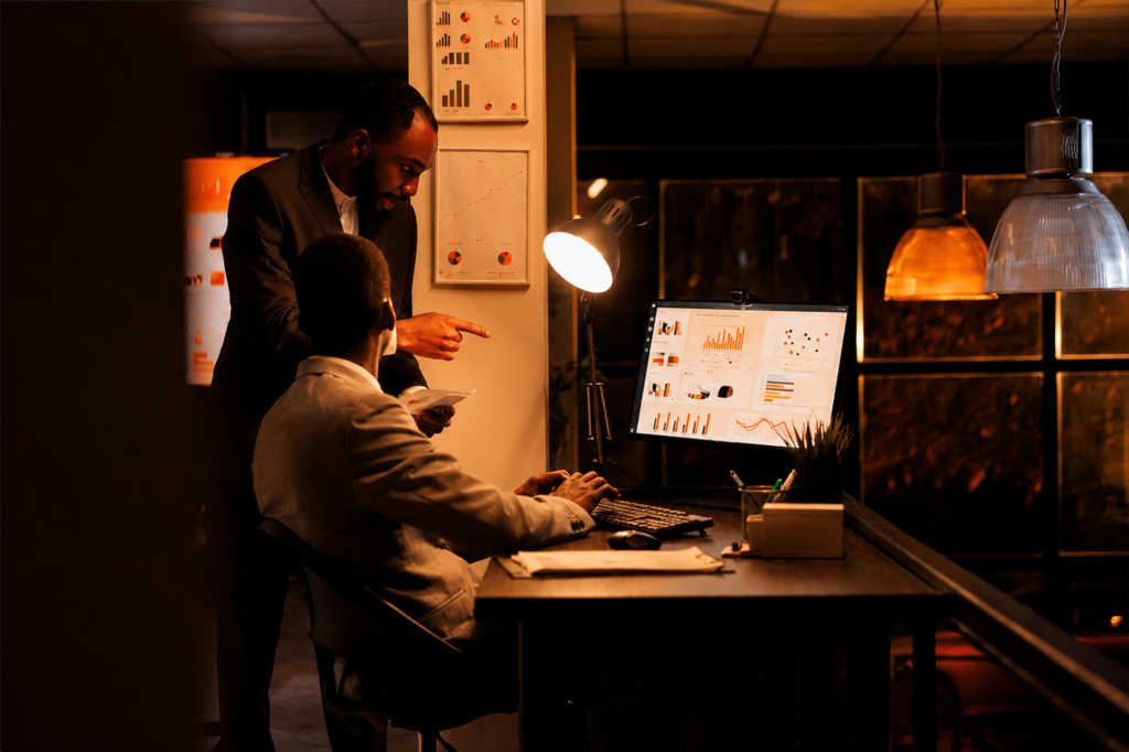 Two men in suits review digital marketing analytics on a computer in a dim office, charts visible on desk and wall.