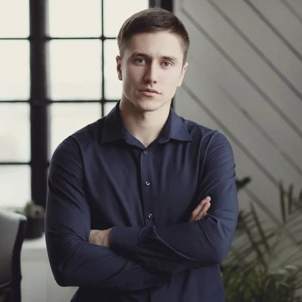 Young man with short hair and dark shirt stands indoors, arms crossed, near a whiteboard on AI automation and web design.